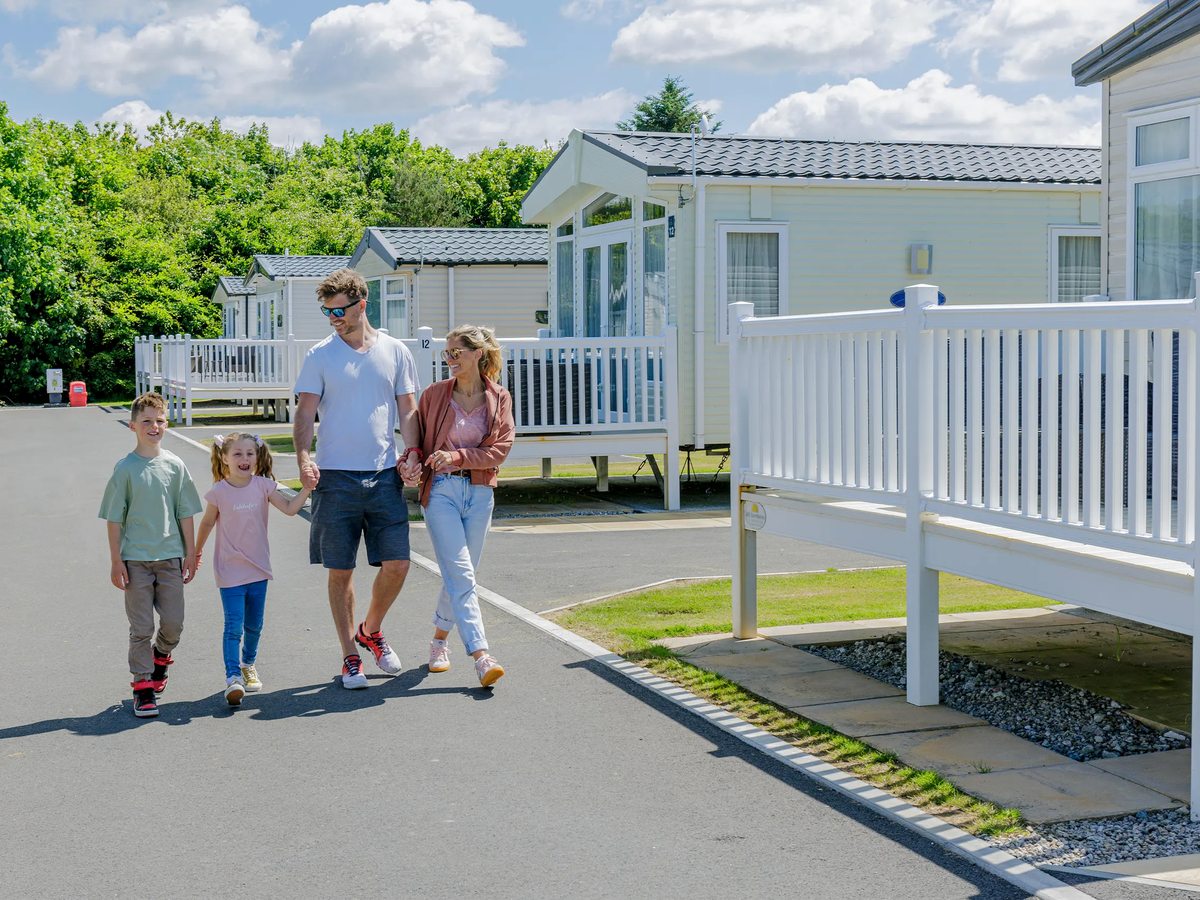 A family walking along a smooth tarmac road lined with cream static caravans on white-railed verandas, trees beyond under a bright sky.