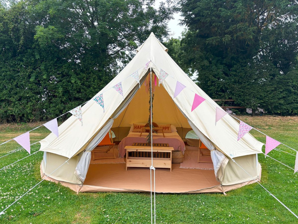 A cream canvas bell tent hung with pastel bunting, its open flap showing a made-up double bed and a wooden storage bench inside, on a mown grass field backed by trees.