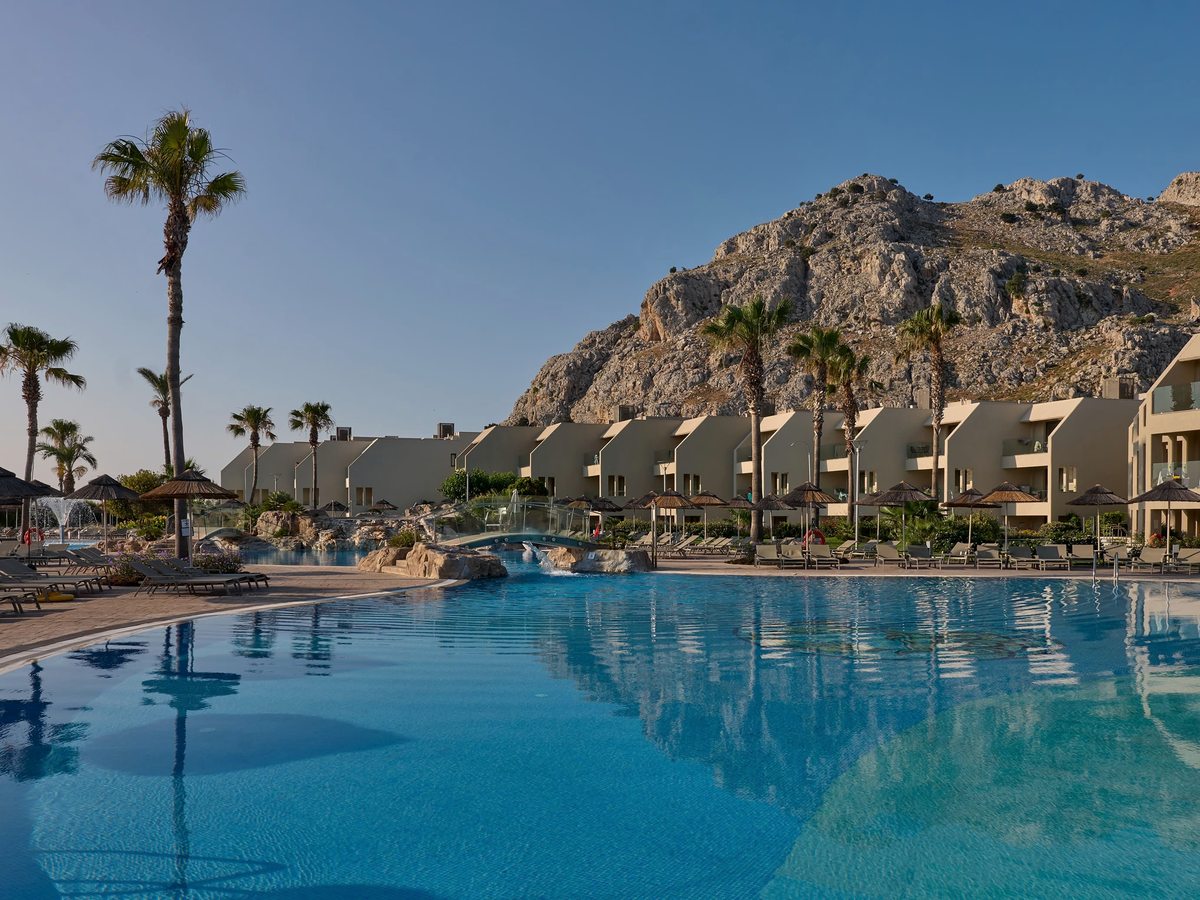 A large curving pool edged with thatched umbrellas and sun loungers, backed by white low-rise buildings and the bare rocky slopes of Tsambika Mountain.