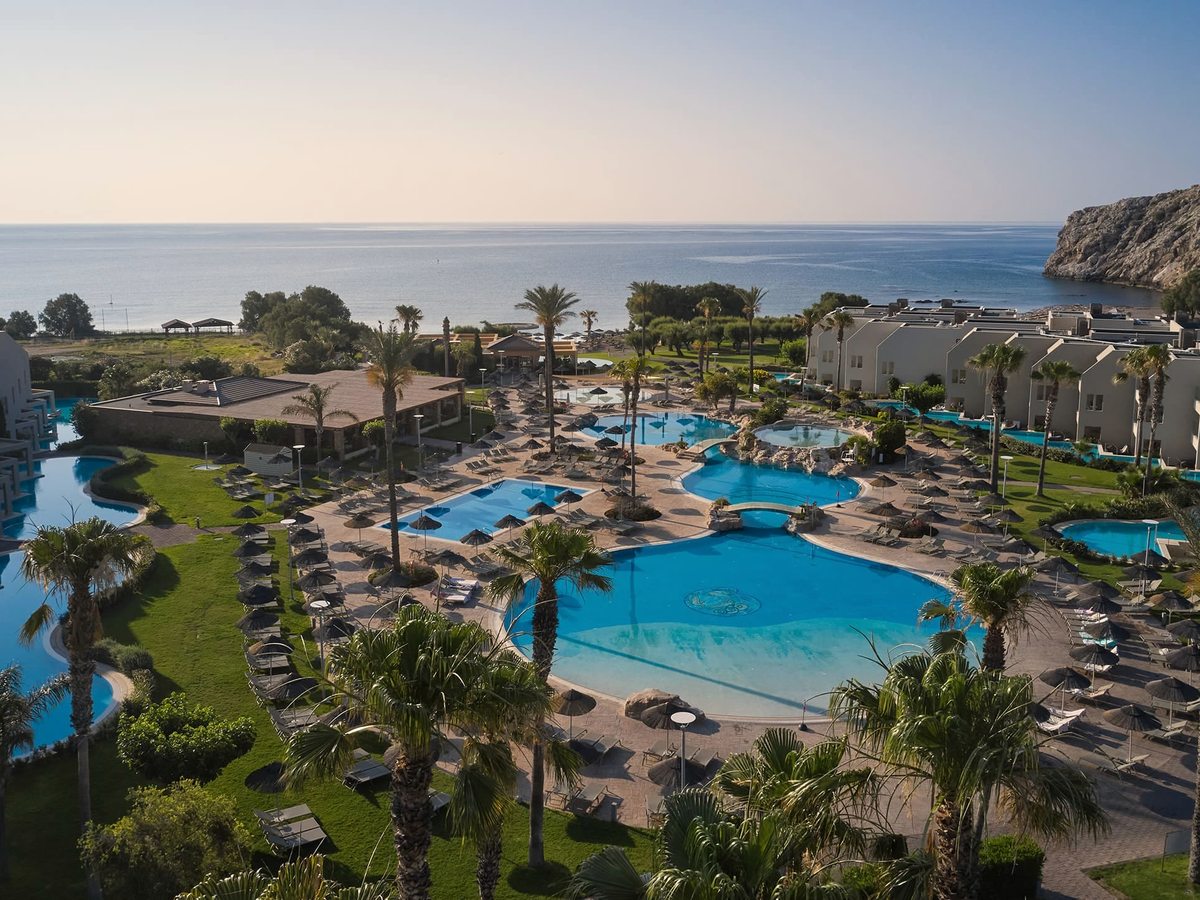 Aerial view of the resort's lagoon pools, thatched umbrellas and palm gardens, with white low-rise buildings, a rocky headland and the sea behind.