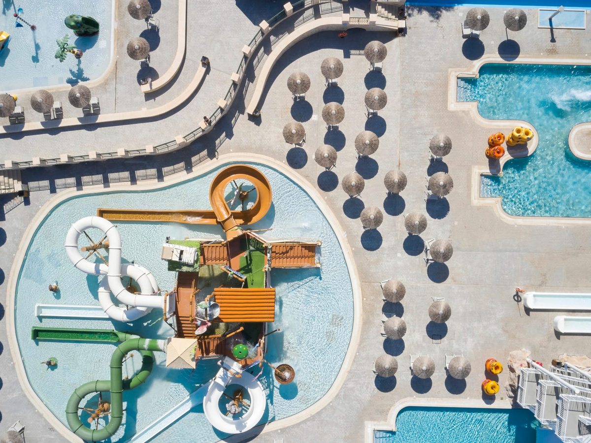 Aerial view of a lagoon-shaped children's play pool with a multi-level wooden play fort, small tube slides and tipping features, ringed by thatched umbrellas.