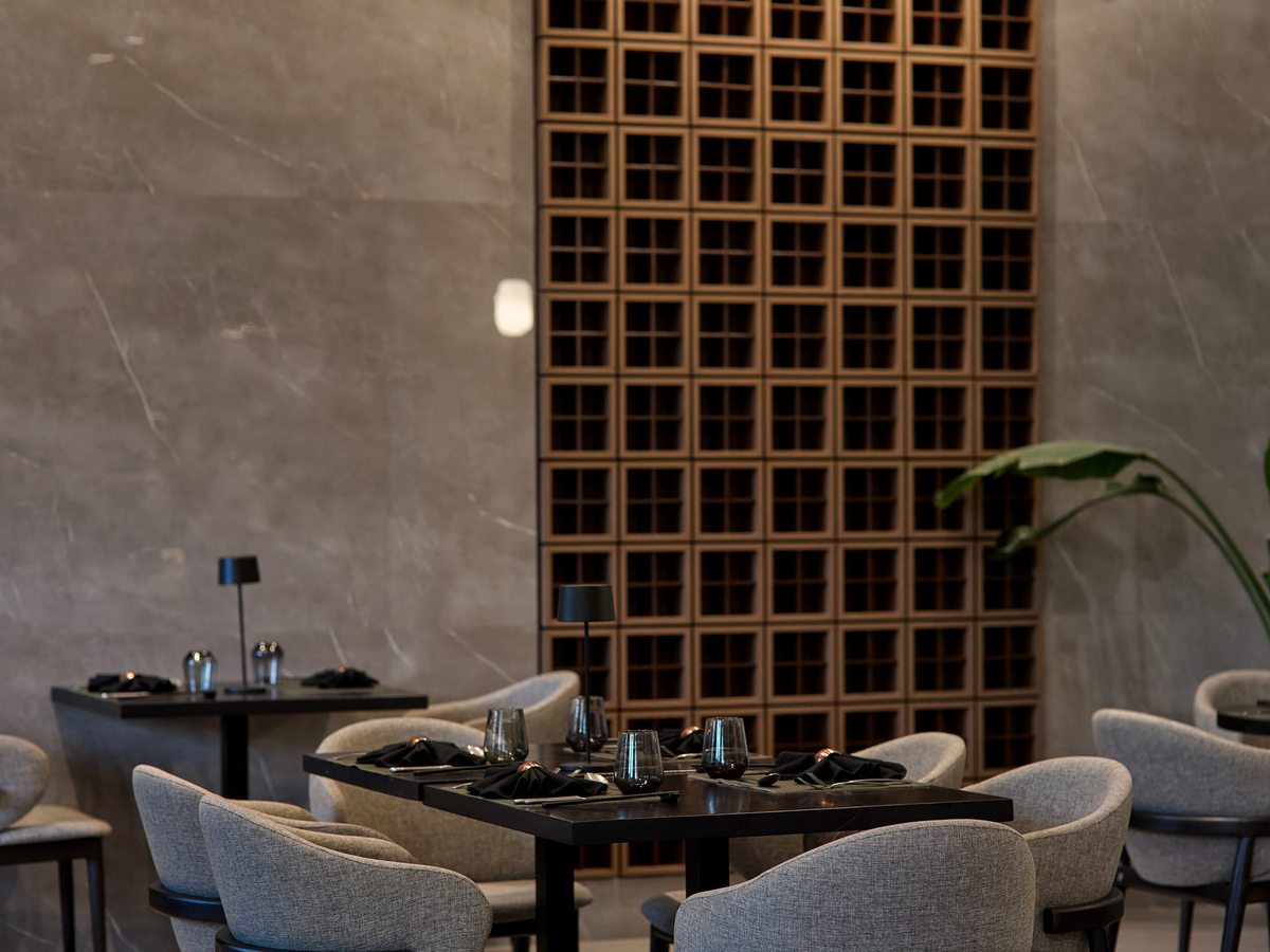 An indoor restaurant with black tables set for dinner, grey tub chairs and a tall latticed timber screen against stone-grey walls.