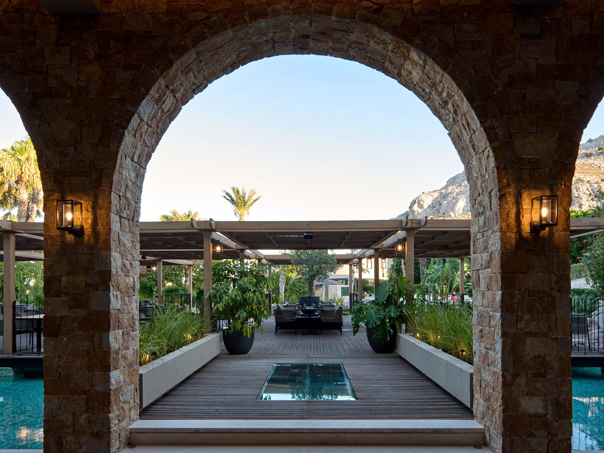 A stone archway framing a timber deck walkway lined with planters and pergolas, leading through the gardens with the rocky mountain beyond.