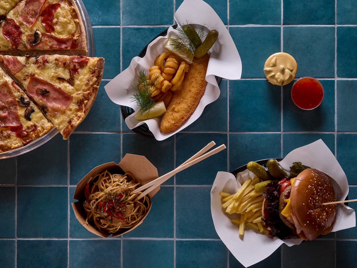 Overhead view of pizza, a burger and chips, and a noodle box on a blue-tiled table.