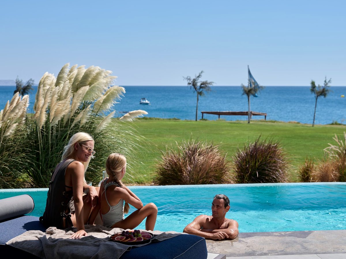 A family sitting at the edge of an infinity pool whose water meets a lawn and the blue Aegean beyond, with pampas grass and a Greek flag.