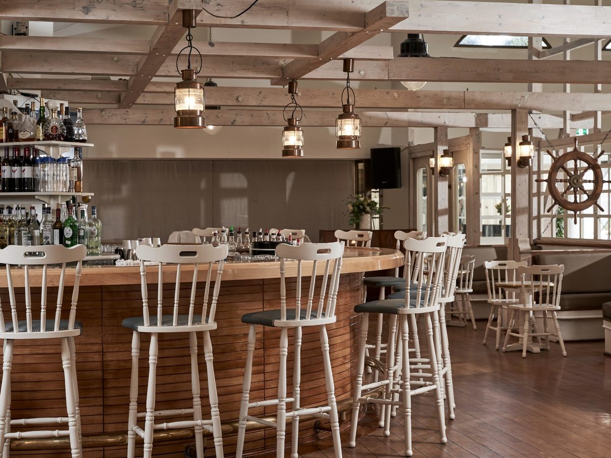 Indoor bar with a curved timber counter and white spindle-back stools, a ship's wheel and hanging lanterns among exposed wooden beams.