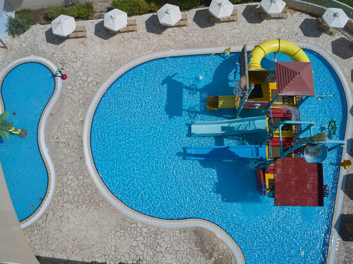 Overhead view of a children's pool with a colourful water-play structure and small slides, and a separate shallow paddling pool beside it, ringed by parasols.