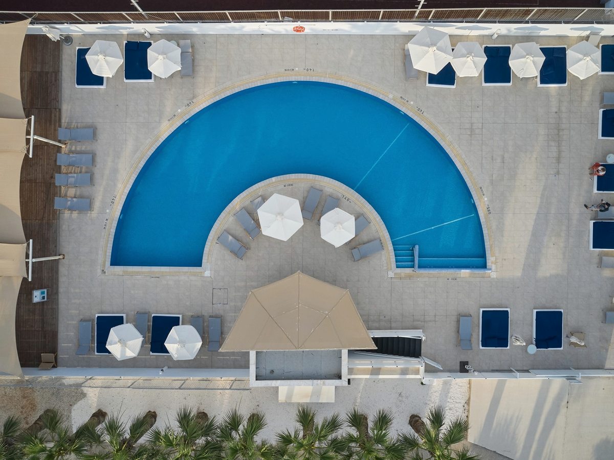 Overhead view of a semi-circular pool with blue sun loungers and white parasols on a paved deck, a shaded seating area at its curved edge.
