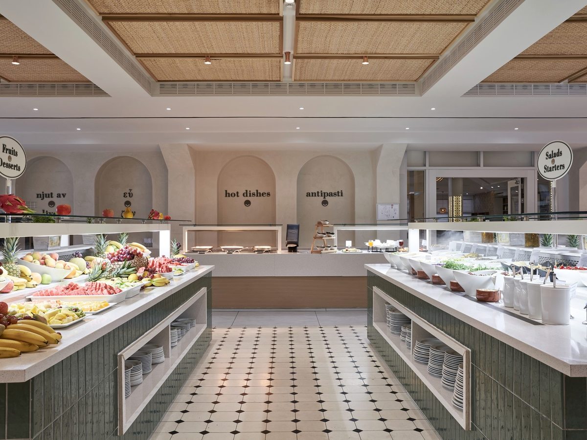 Buffet hall with hot and cold serving counters, a fresh-fruit display and stacked plates along a wide tiled aisle.