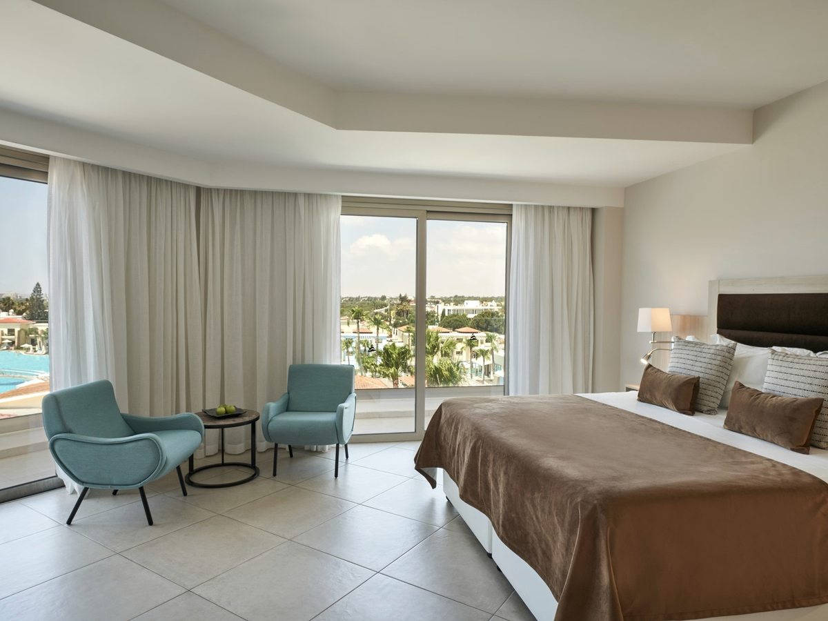 Bedroom with a double bed and a separate seating area of blue armchairs by wide windows, looking out over the pool and resort gardens.