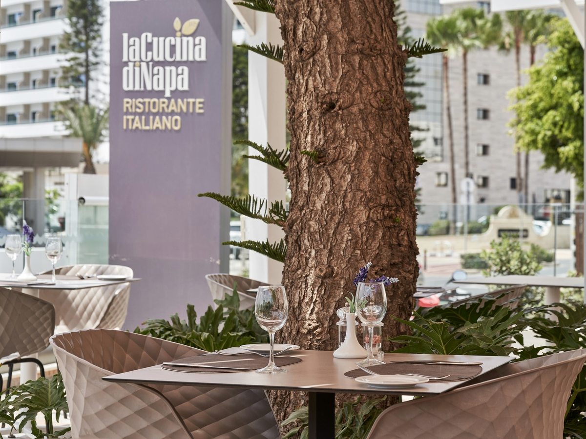 Outdoor Italian restaurant terrace with tables laid under mature trees, a sign reading La Cucina di Napa Ristorante Italiano behind.