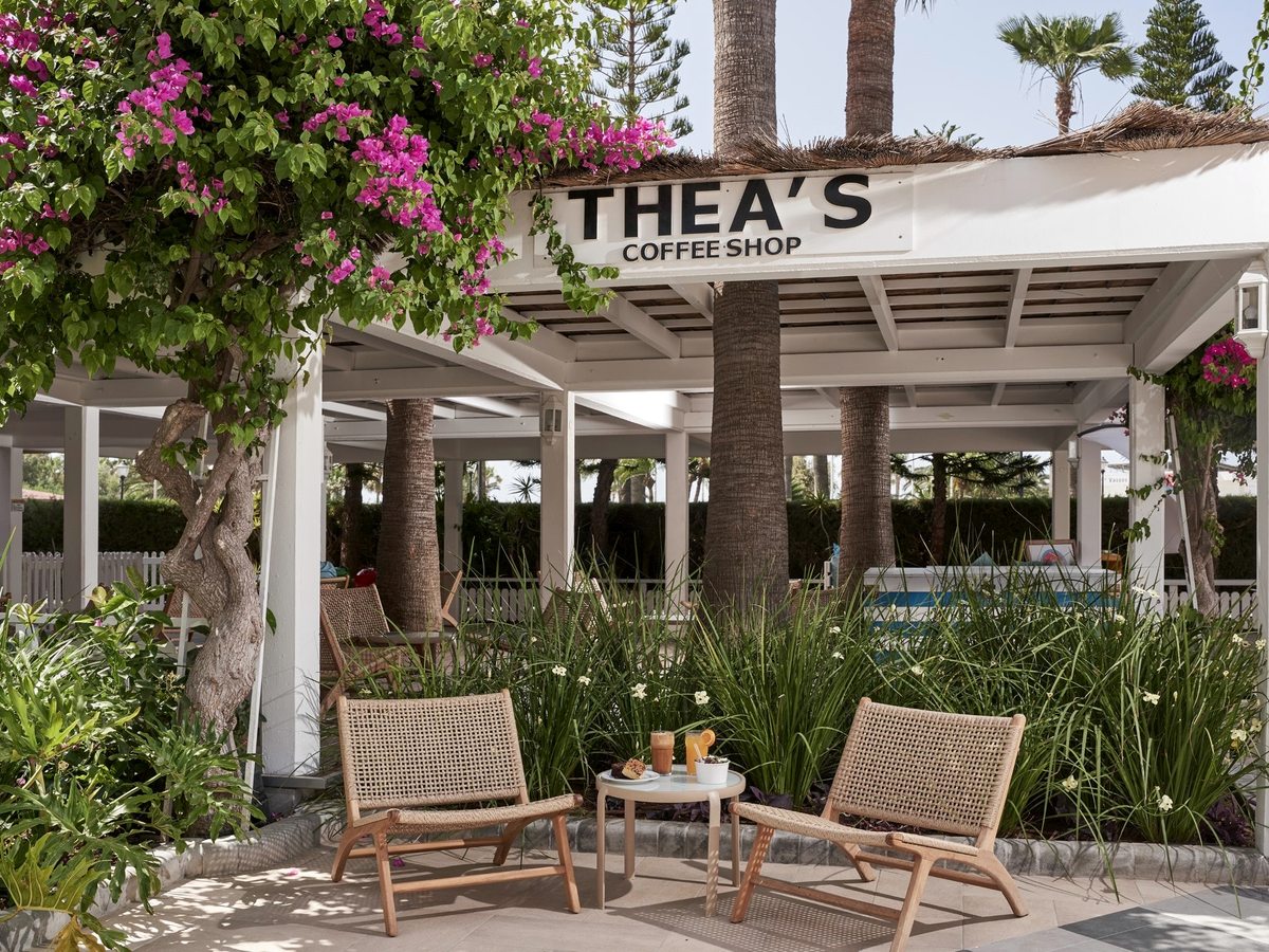 Open-sided garden coffee bar under a white pergola with wicker armchairs, framed by palms and pink bougainvillea, a Thea's Coffee Shop sign above.