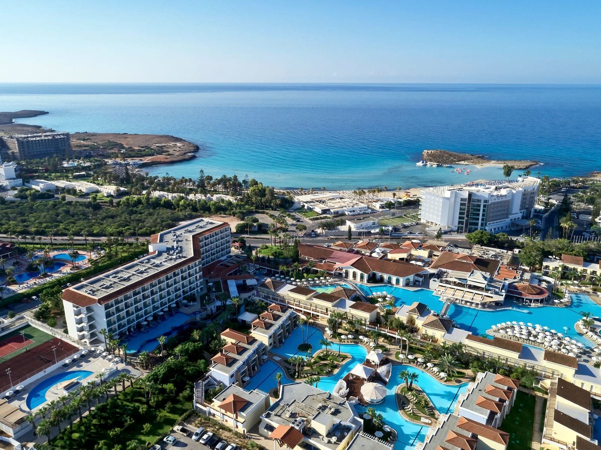 Aerial view of the resort by the sea, turquoise lagoon pools among low terracotta-roofed buildings and palm gardens, with a sandy bay and a small islet beyond.