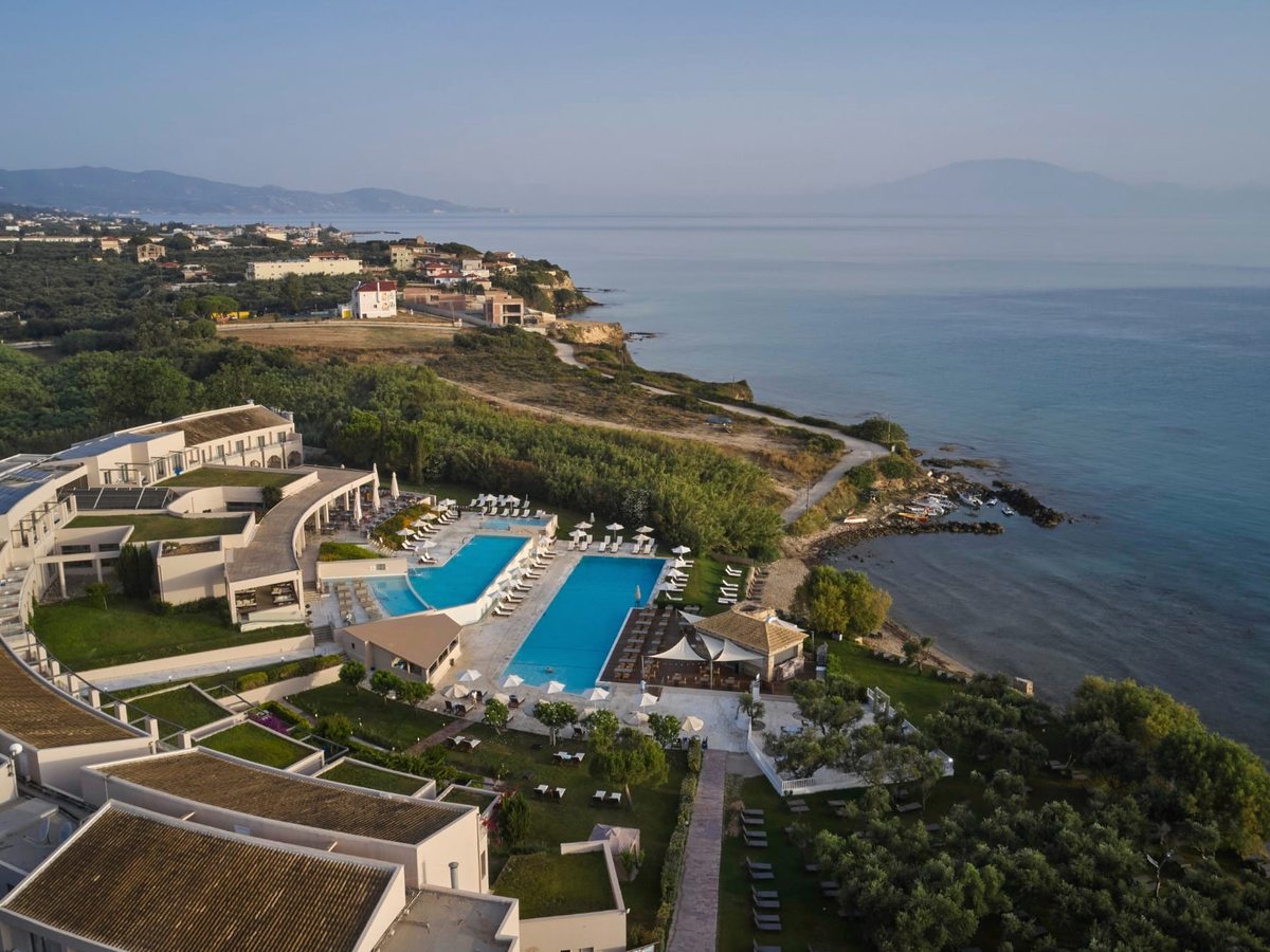 Aerial along the coastline showing the resort and its pool above a rocky, sandy shore, with the sea and distant mountains beyond.