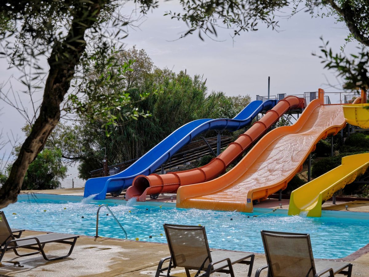 Four coloured waterslides, blue, red, orange and yellow, curving down into a shallow landing pool beside sun loungers.