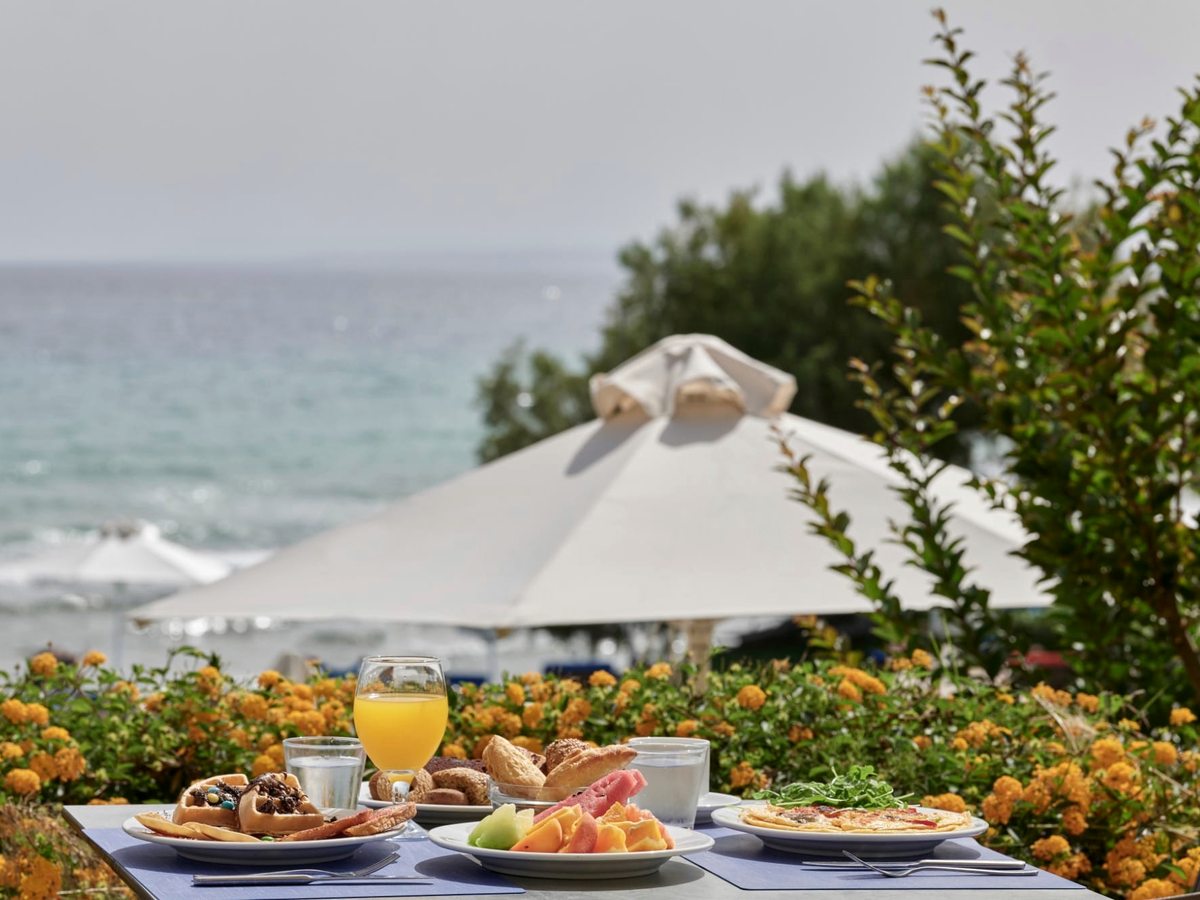 A table set with pastries, fruit and juice under a parasol, flowering plants in front and the sea behind.