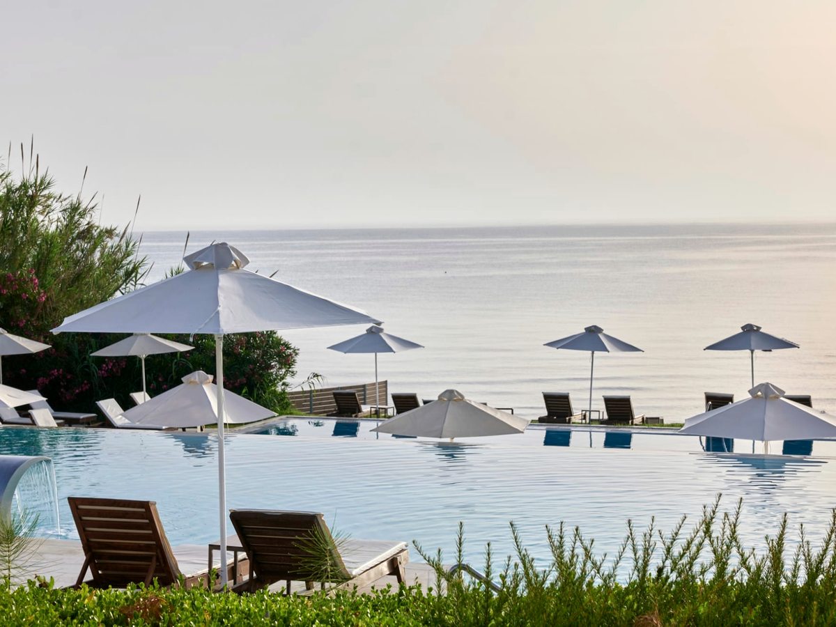Outdoor pool with white parasols and loungers along a planted edge, the sea filling the horizon beyond.