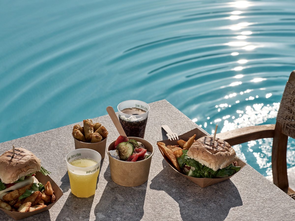 A burger, chips, salad and a glass of juice on a table at the edge of the pool in bright sun.