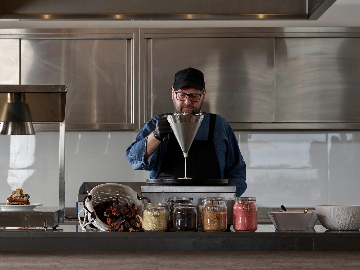 A chef in a blue shirt and black apron preparing food at a steel show-cooking counter lined with jars and garnishes.