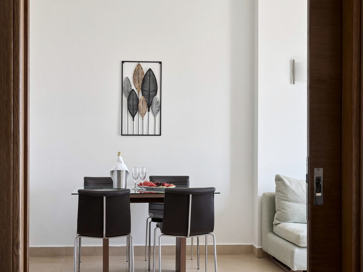 A dining table with dark chairs and a sparkling-wine bucket in a suite, with the bedroom visible through a doorway beyond.