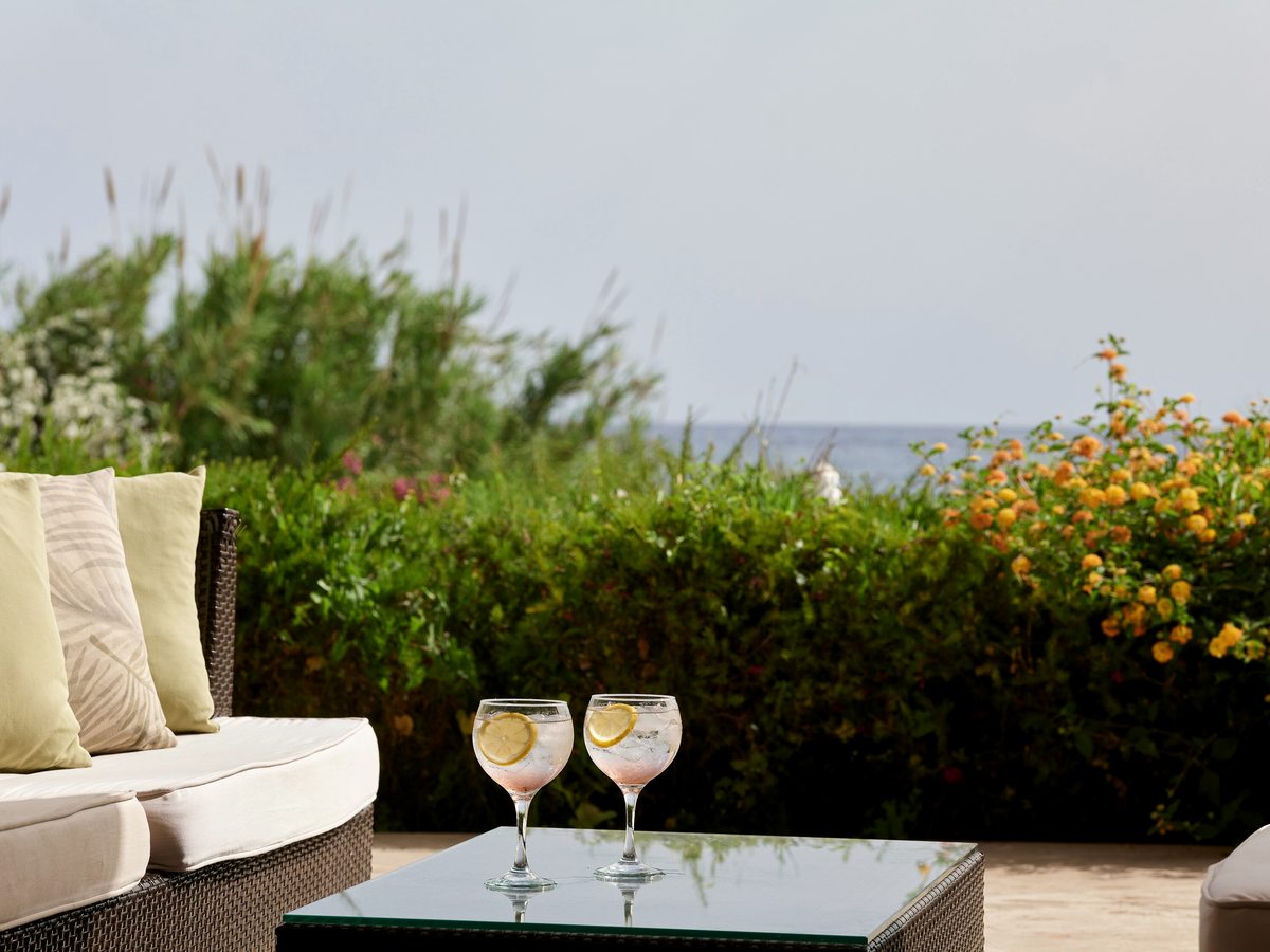 Cushioned wicker sofa with two glasses on a low glass table, a flowering hedge in front and the sea beyond.