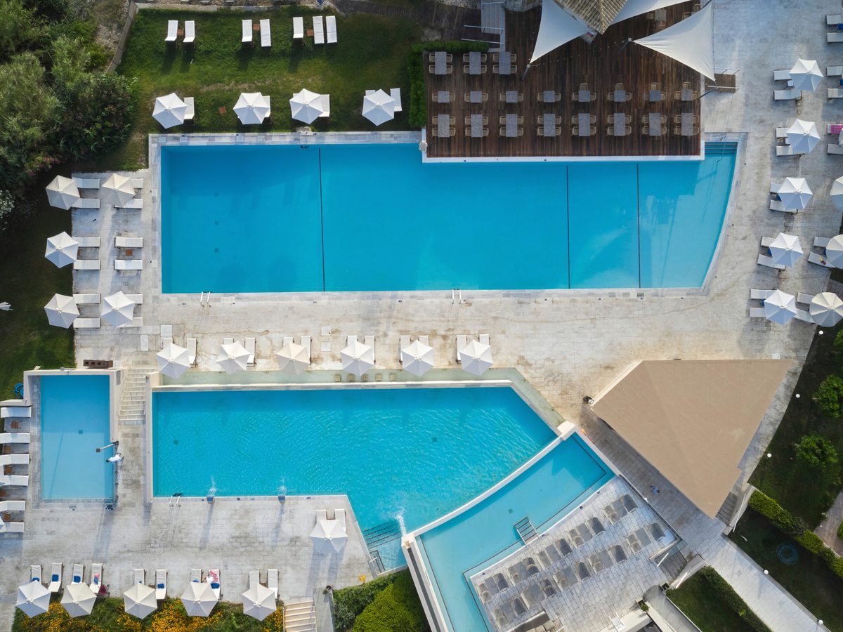 Aerial of two linked blue swimming pools edged by rows of white loungers and parasols on a paved deck.