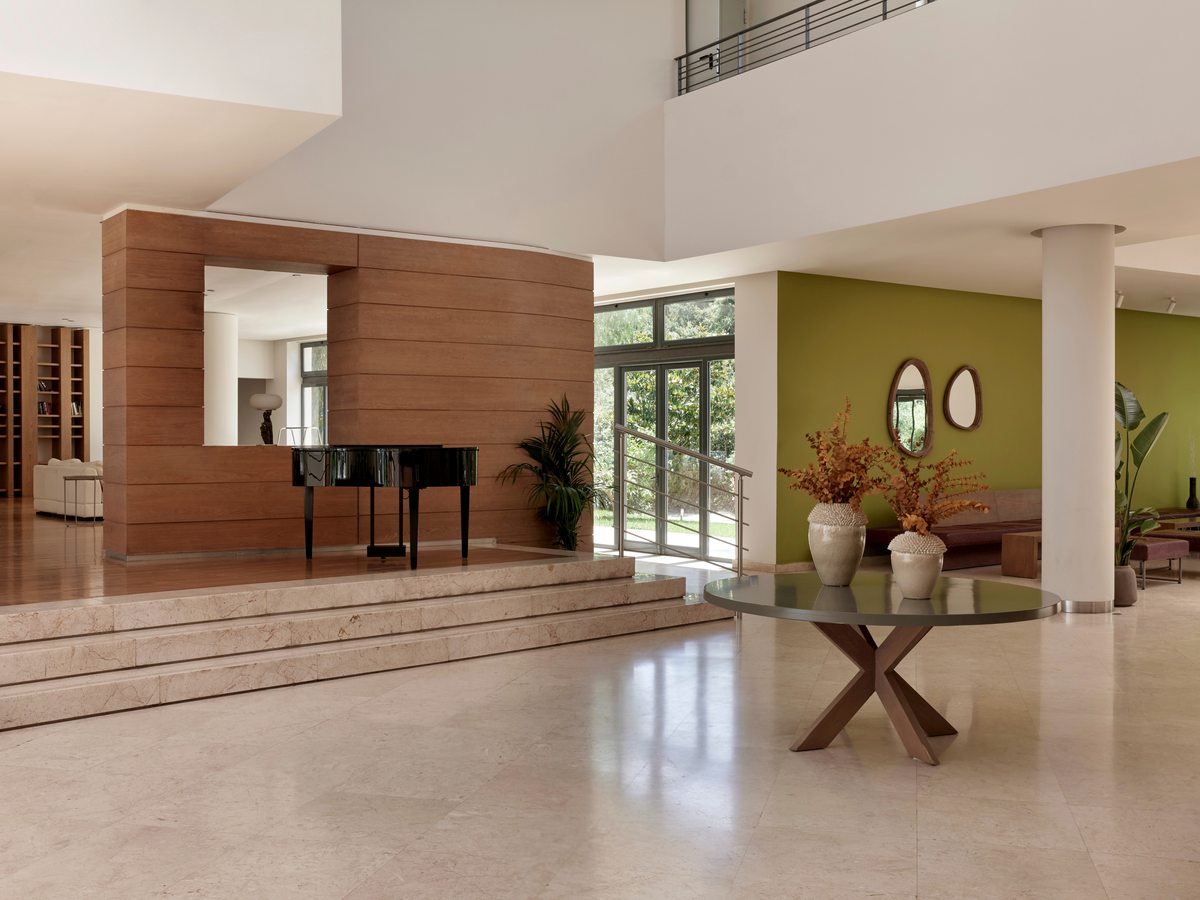 Double-height lobby with a grand piano, broad stone steps, white columns and timber wall panelling over a polished stone floor.