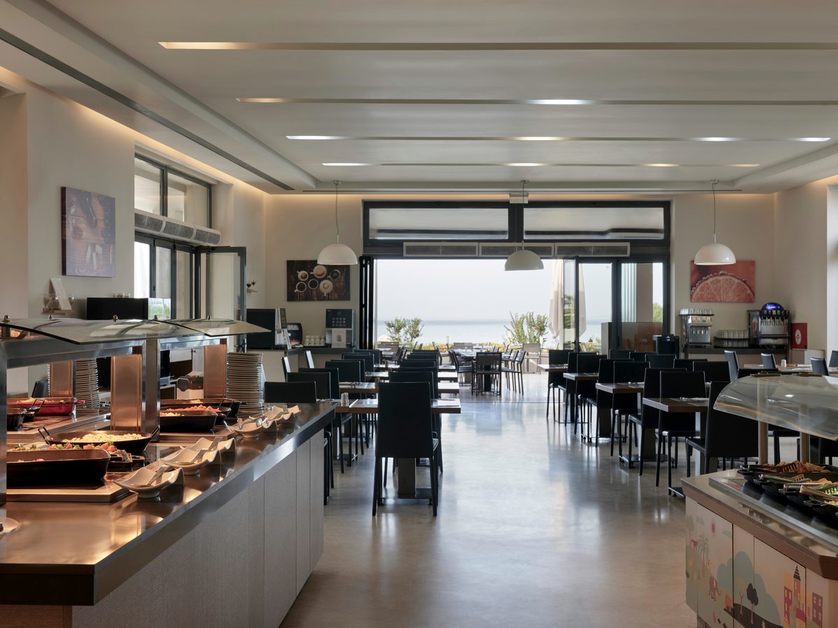 Long buffet counter with hot-food stations running down a bright dining hall of dark chairs, with sea-view windows at the far end.