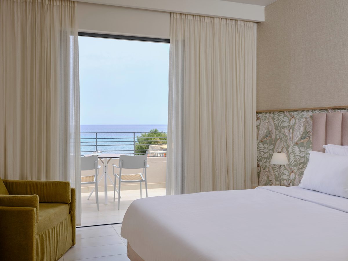 Double room with a white bed and upholstered headboard, a mustard sofa and glass doors to a terrace with a sea view.