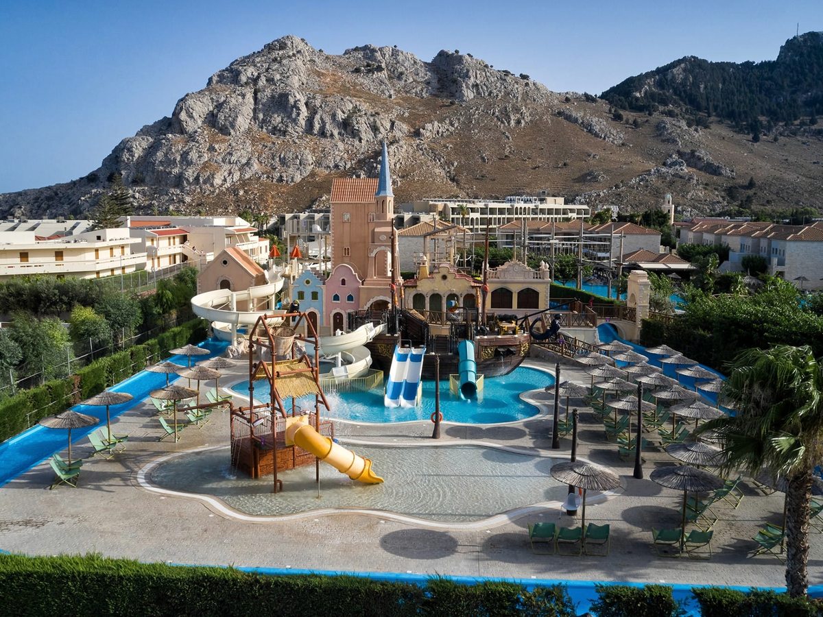 Children's waterpark with a central pirate-themed play structure, small slides and a shallow pool ringed by loungers, mountains behind.