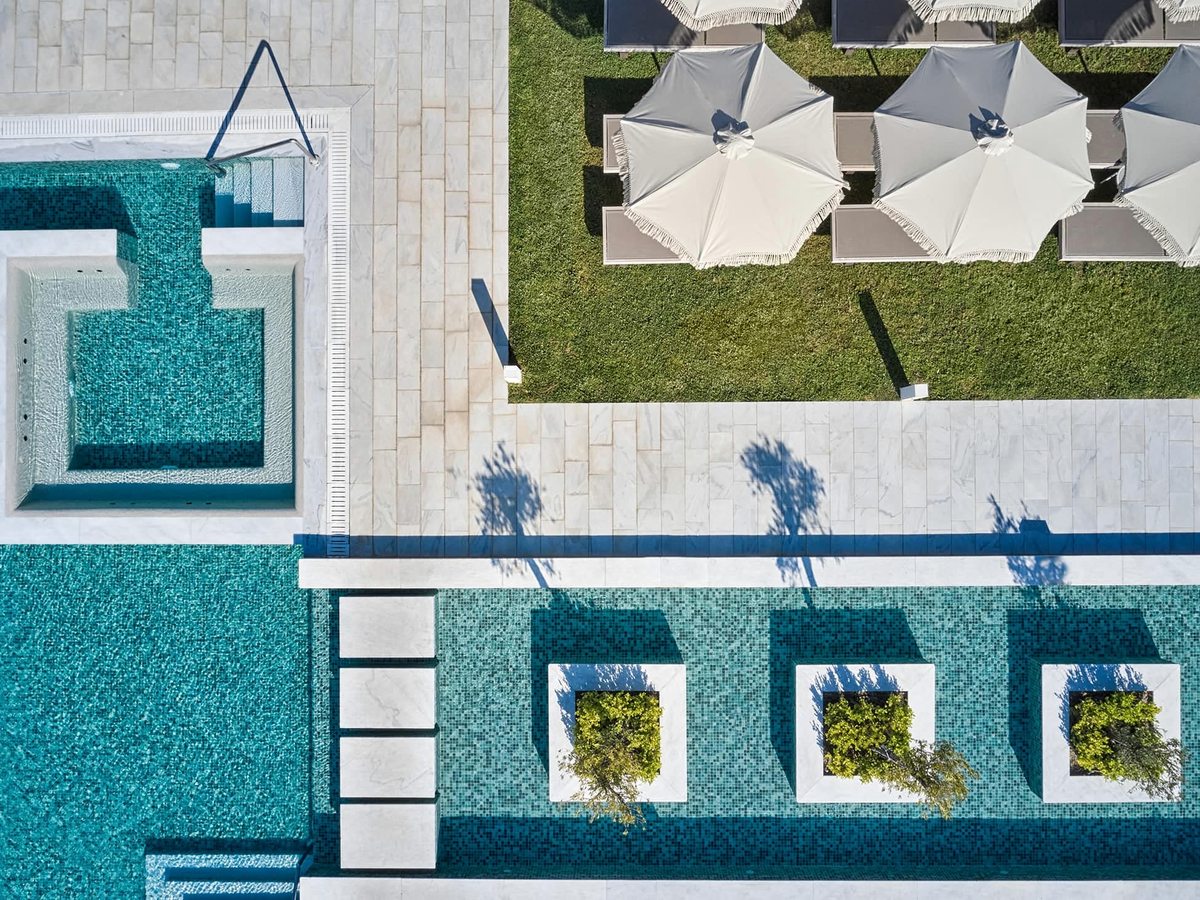 Aerial view of a rectangular pool with square planted islands and a stepping-stone walkway, white paving, parasols and loungers.
