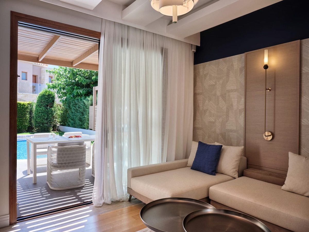 Suite lounge with a cream corner sofa and a daybed with a blue cushion against a wood-panelled wall, sheer curtains and sliding doors to a terrace.