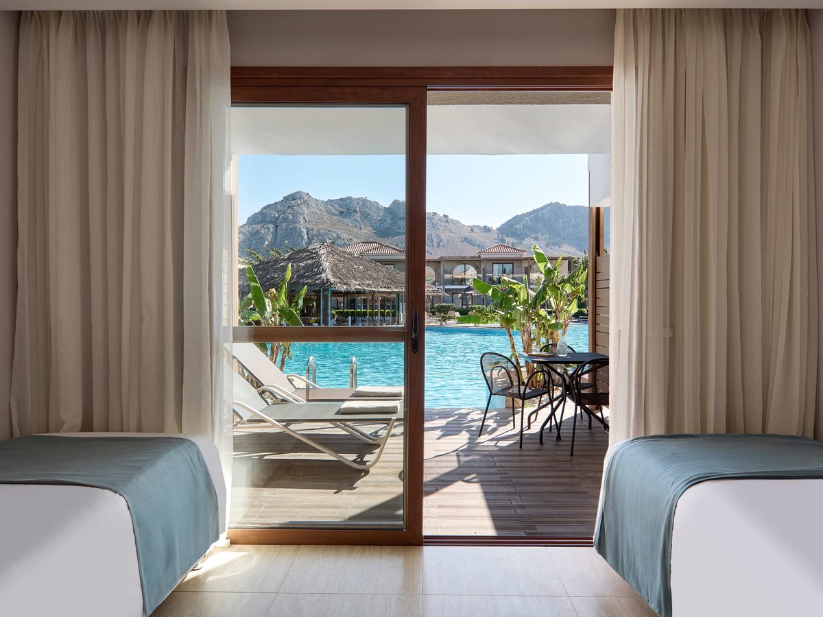Twin room looking through sliding doors onto a decked terrace with loungers and a shared pool directly outside, mountains behind.