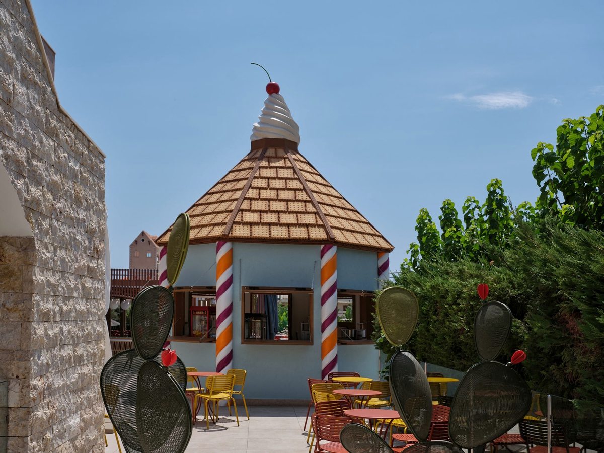 Small outdoor kiosk with an ice-cream-cone roof and candy-stripe columns, ringed by yellow chairs, red tables and cactus planters.