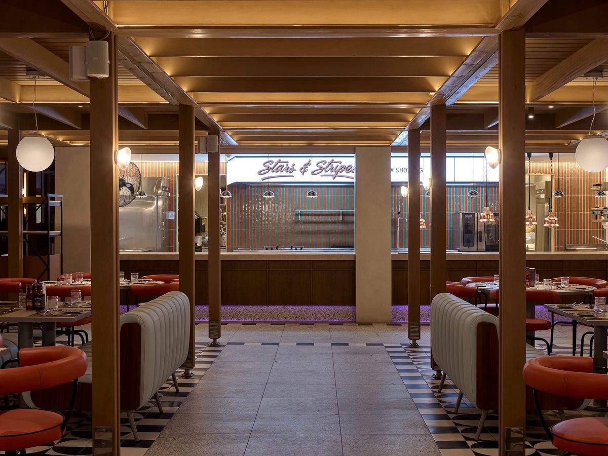 American diner with red booth seating, round tables, a chequerboard floor, timber beams and a service counter with signage at the back.