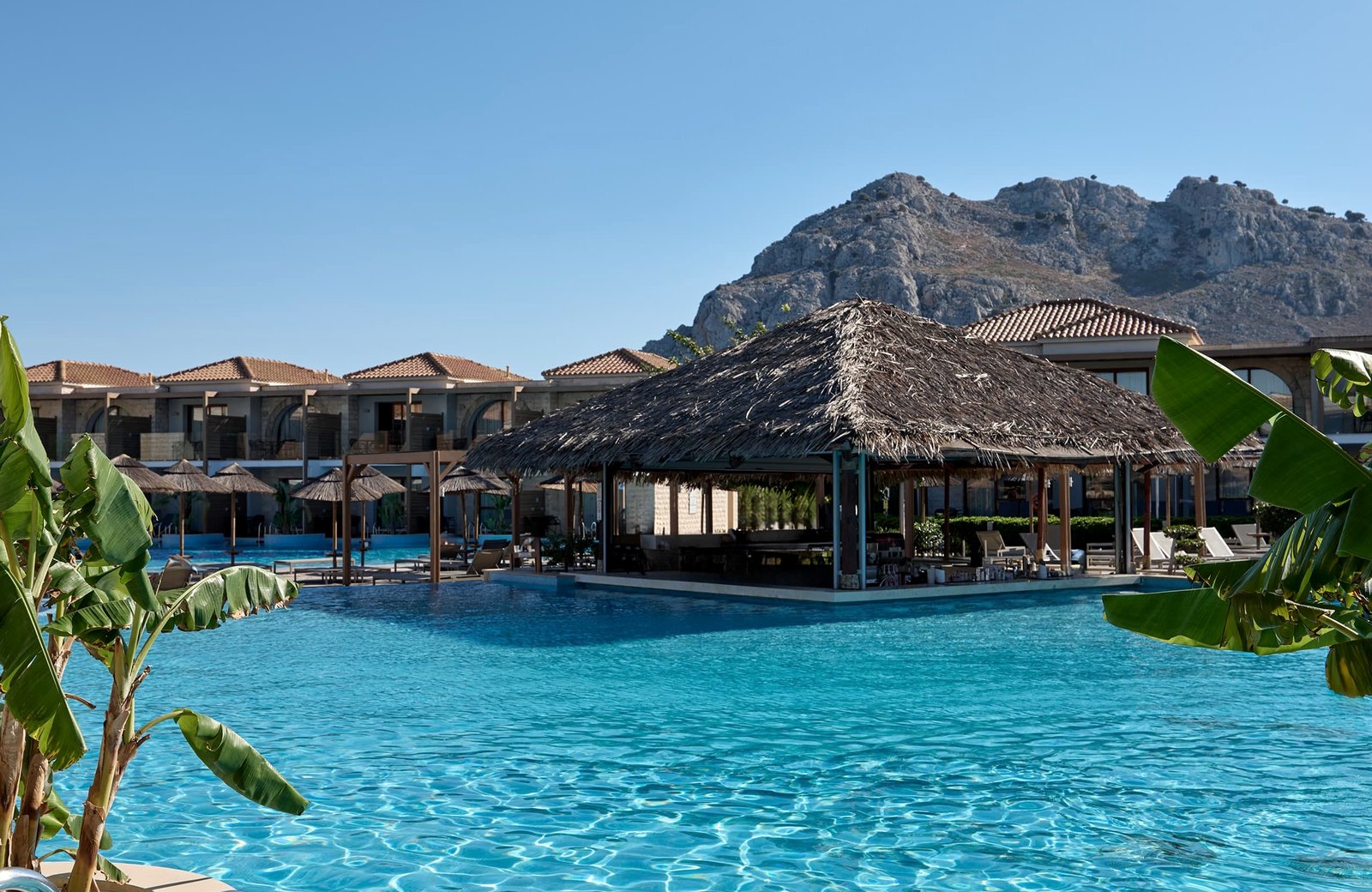 Atlantica Holiday Village Rhodes: the main pool and its thatched bar below Mount Tsambika, with low-rise rooms around it.