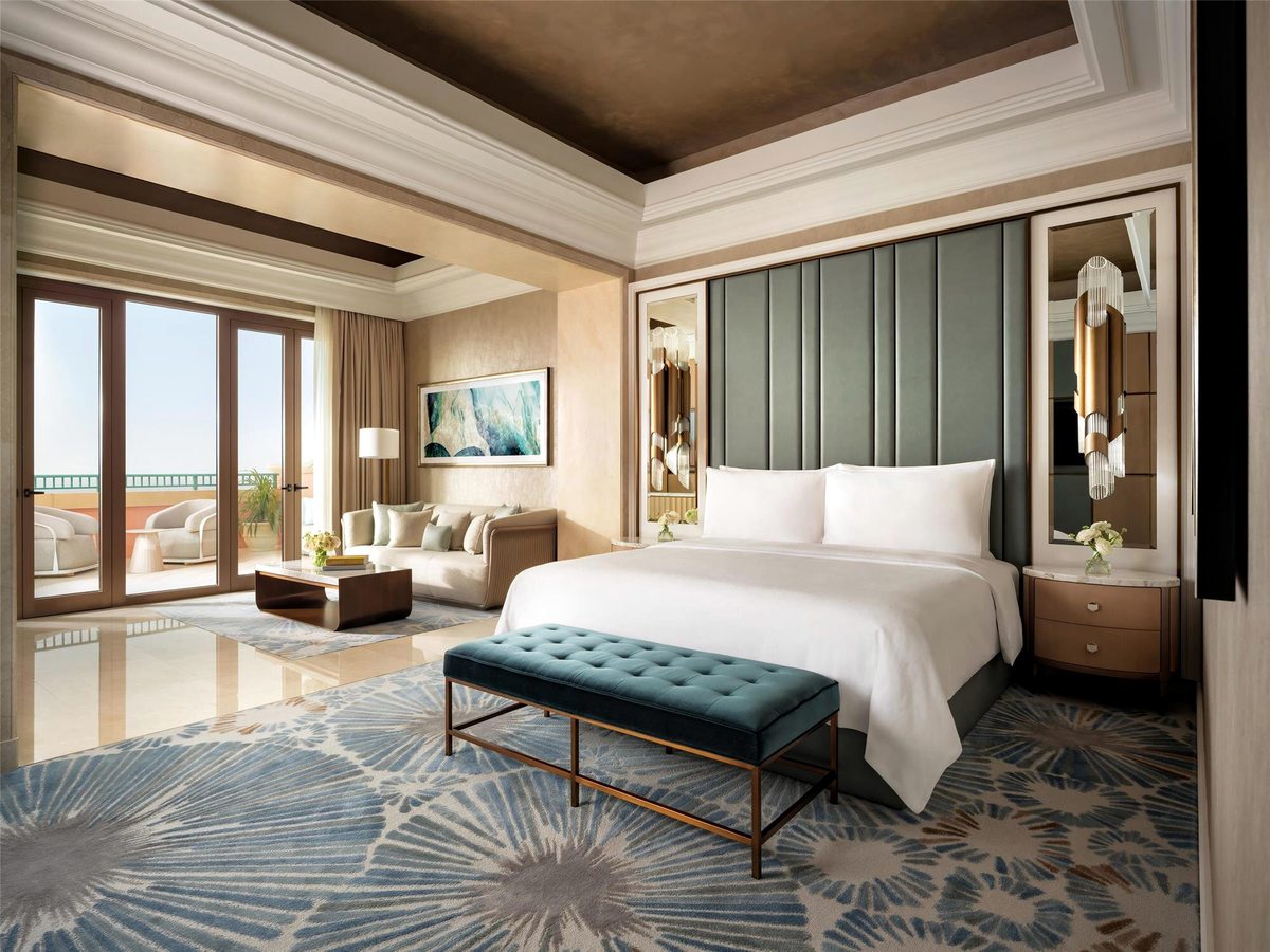 A spacious suite bedroom with a king bed and padded green headboard, a sofa and armchairs by a balcony with a sea view, and a starburst-patterned carpet.