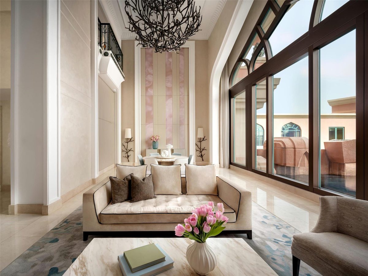 A tall, light-filled suite lounge with a cream sofa, a marble coffee table and a small dining table by soaring arched windows that look onto a terrace and the pink resort facade.