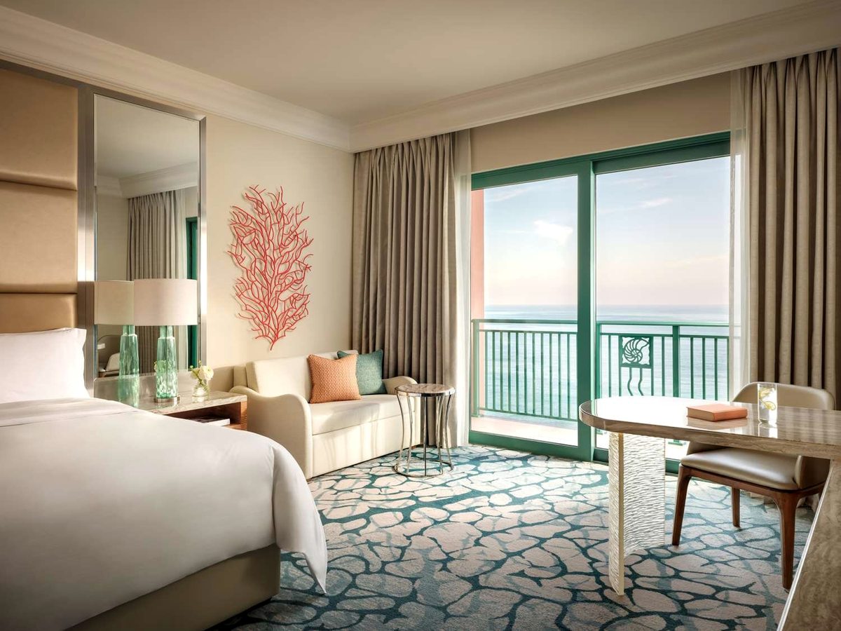 A guest room with a king bed, a cream sofa, a round writing table and a balcony with an open sea view, coral-branch wall art and a coral-patterned carpet.