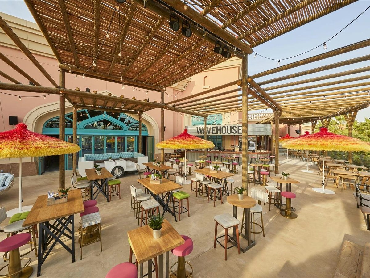 An open-air restaurant terrace under a bamboo canopy strung with lights, with wooden high tables, colourful stools and thatched parasols, the Wavehouse sign on the building behind.