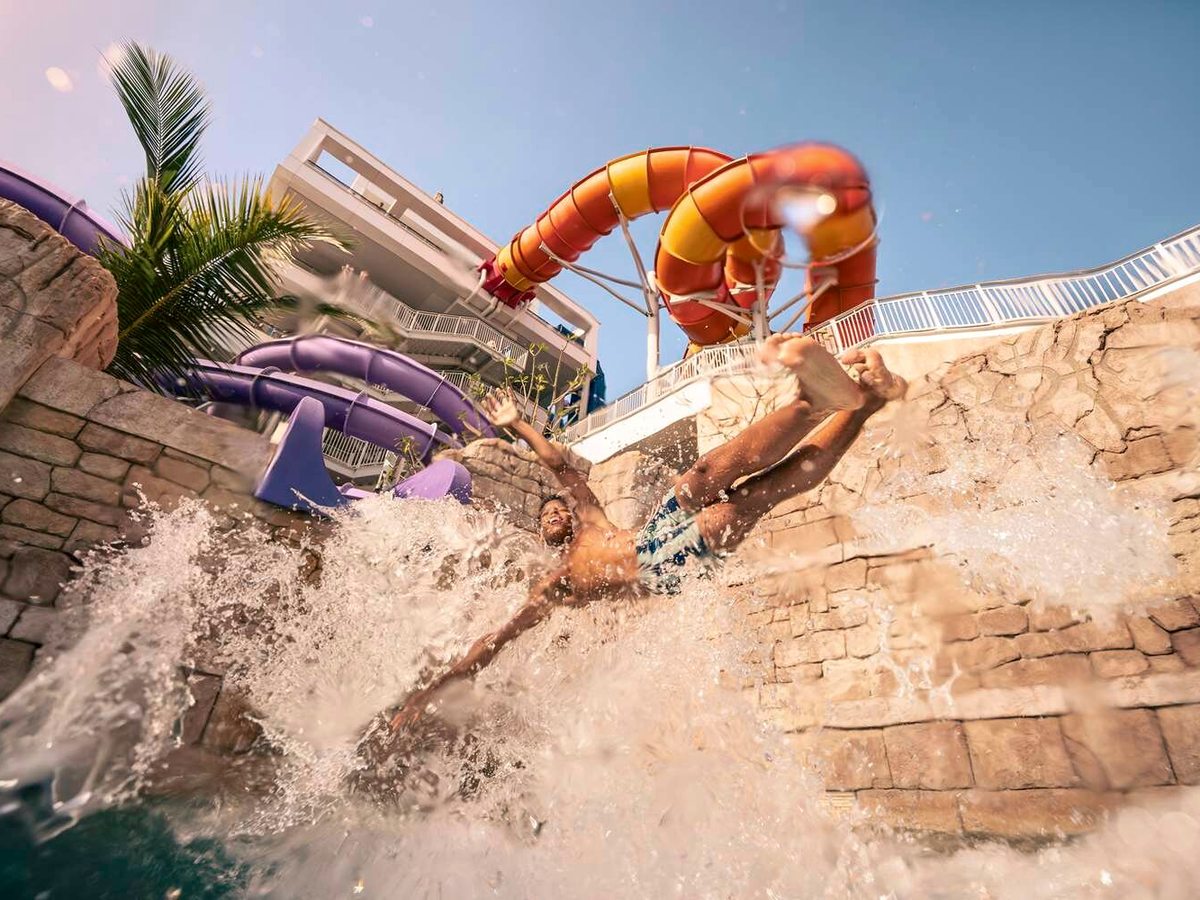 A person splashing down into the pool at the foot of a stone slide tower, with looping orange and purple waterslides above against a blue sky.
