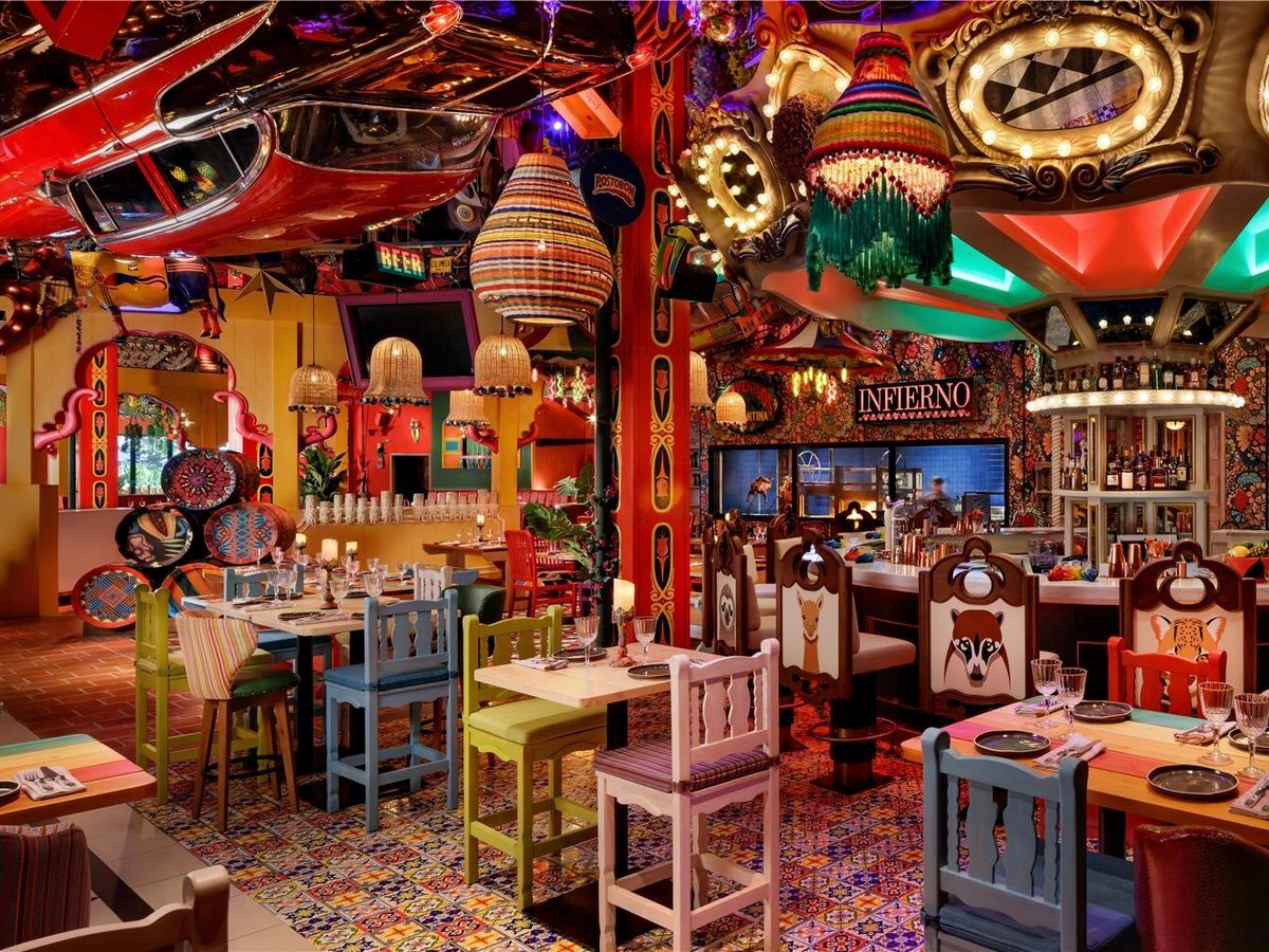 A vividly decorated restaurant with painted animal-motif chairs, a patterned tiled floor, colourful woven lanterns and a vintage car mounted overhead, with an open bar and grill counter.