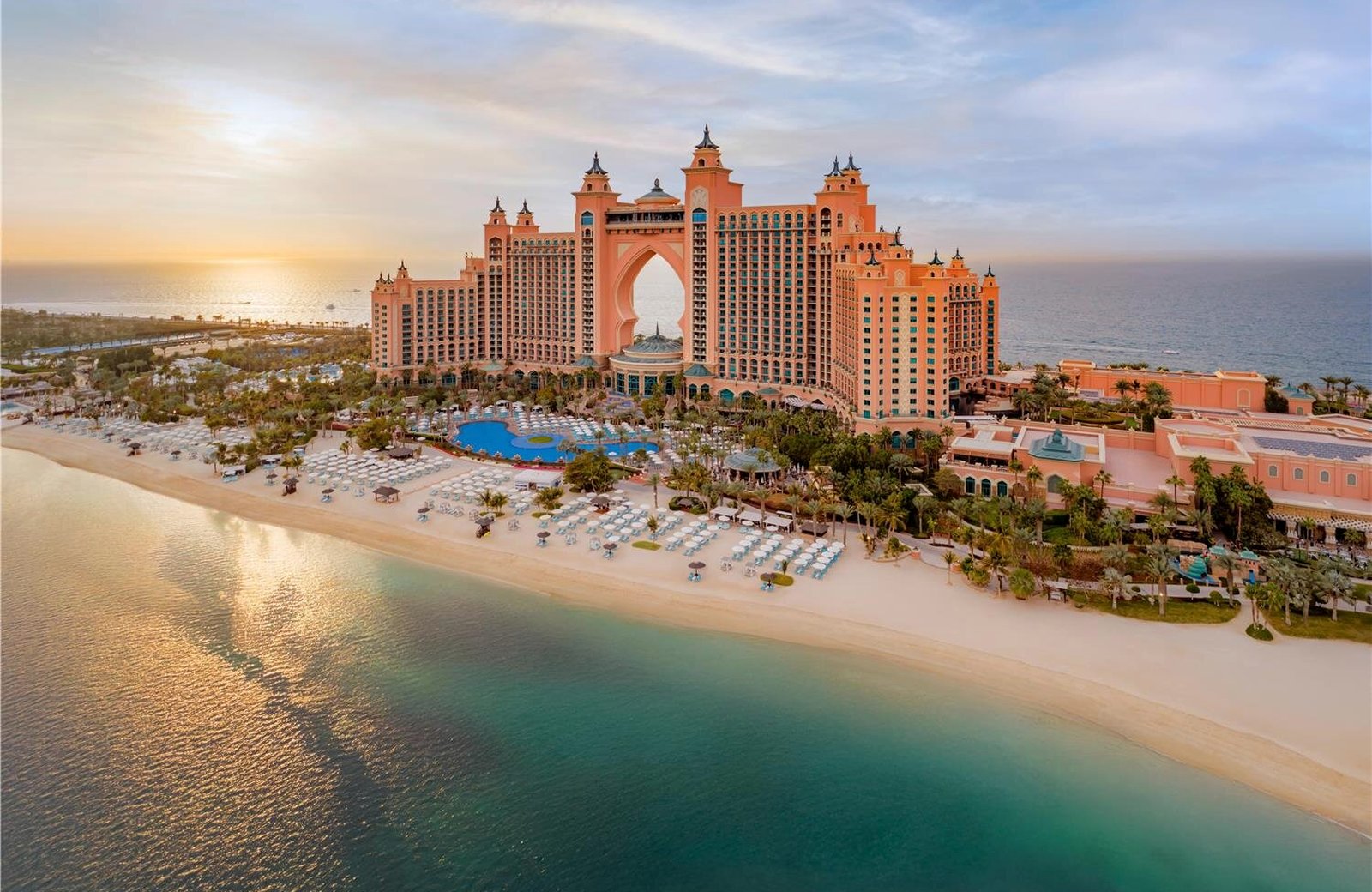 Atlantis, The Palm seen from the air at sunset: the salmon-pink twin-towered resort at the tip of the Palm Jumeirah in Dubai, its central arch above the grounds, with the beach and the Arabian Gulf in front.