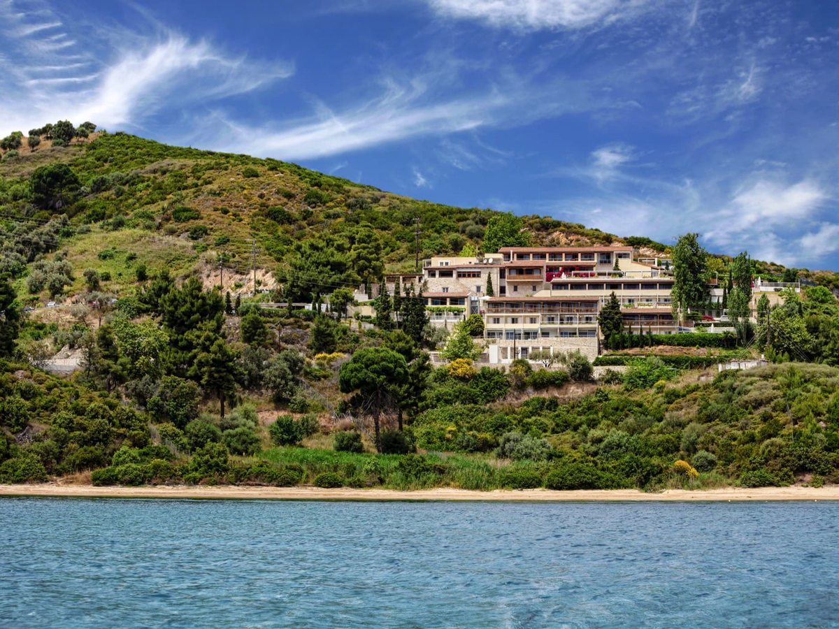 A calm blue bay and a strip of sandy beach at the shoreline, the hotel's low stone buildings stepping up a pine-covered hillside above, under a clear sky.