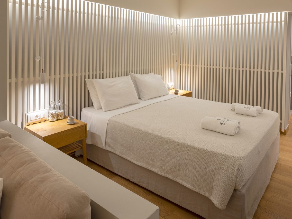 A double room with a low bed, a white slatted wall, a daybed sofa with folded towels and soft lighting over a wood floor.