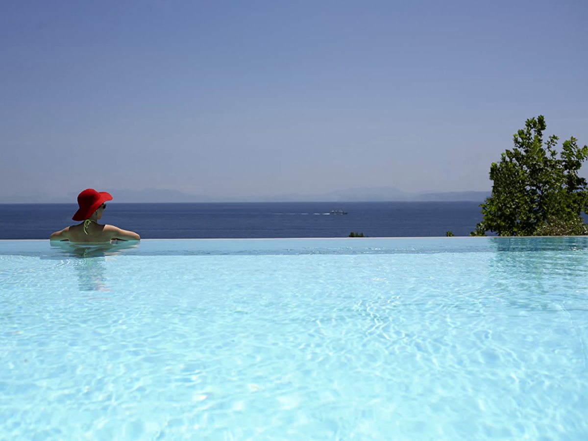 An infinity-edge pool with clear water meeting the horizon, a swimmer in a red sun hat, and open sea and a distant coastline beyond.