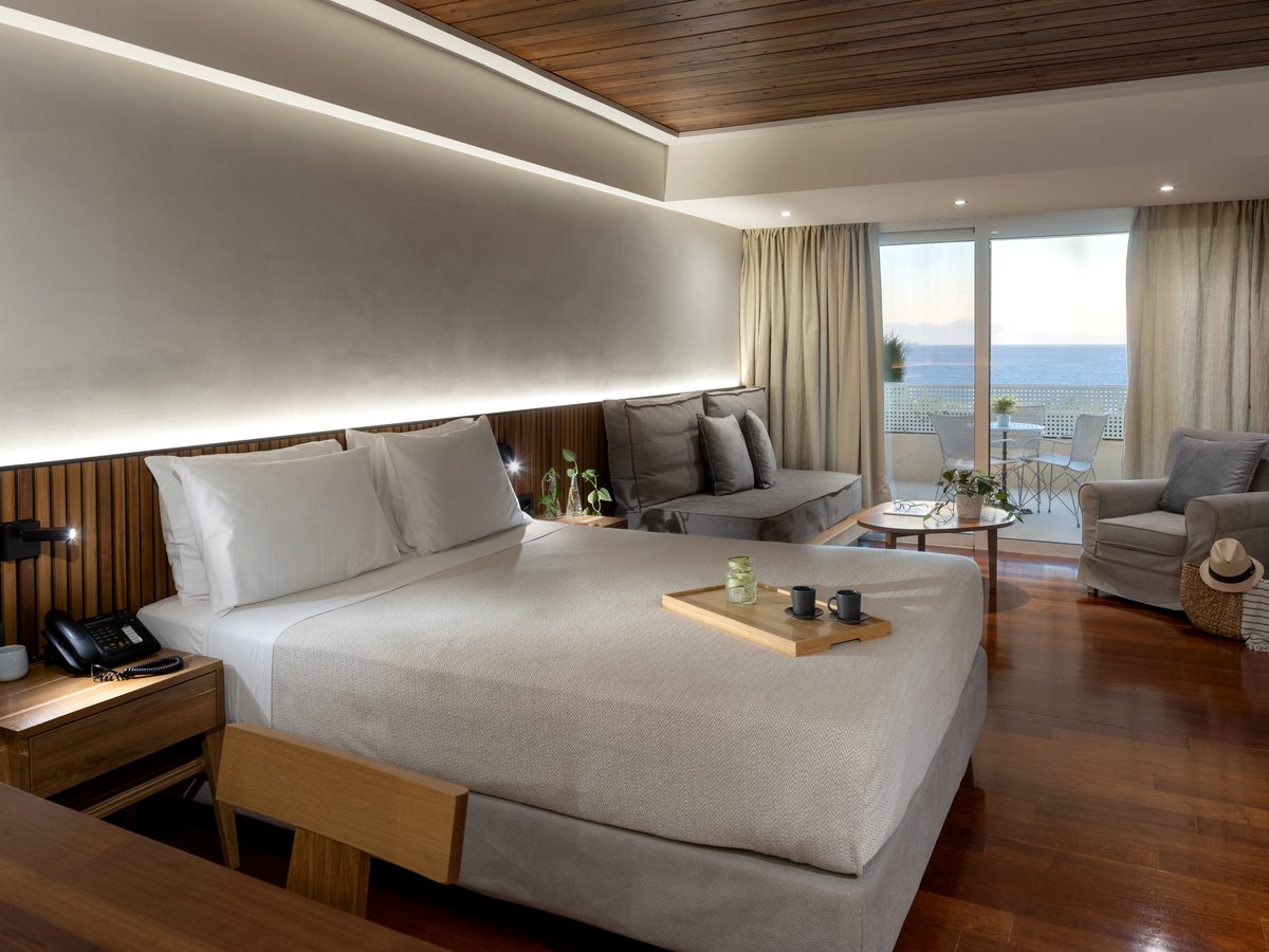 A double room with a wood-panelled headboard, a large bed, a sofa-bed seating corner and balcony doors opening to a sea view over a polished wood floor.