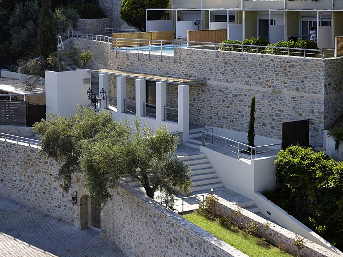 Whitewashed stairways and stone retaining walls stepping down a hillside between accommodation blocks, with an olive tree and a small terrace pool above.