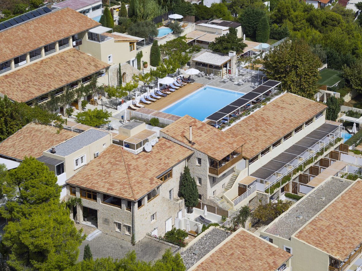 An aerial view of the hotel's terracotta-roofed stone buildings around a rectangular blue swimming pool, on planted garden terraces with the sea beyond.