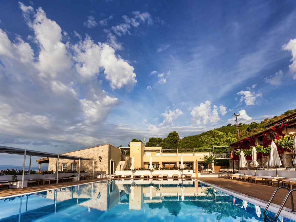 A rectangular swimming pool with entry steps, sun loungers and parasols on the deck, cream hotel buildings behind and a hillside sea view.